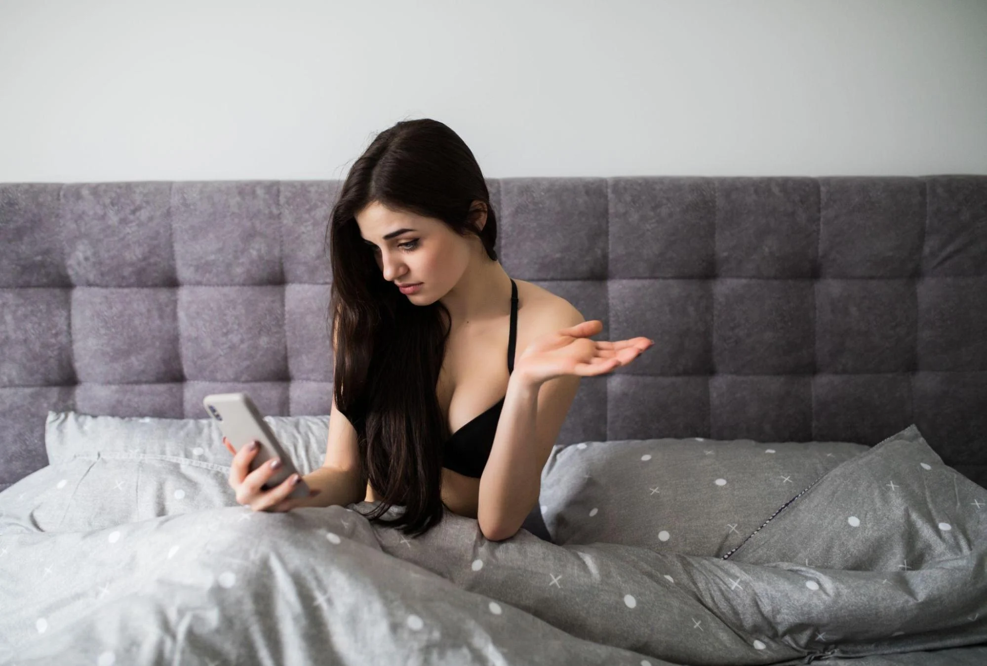 A woman wearing a black bra sits on a bed while holding a smartphone in one hand, appearing to be on a video call.
